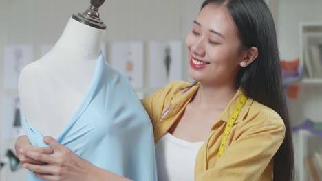 primer plano de una diseñadora asiática sonriente con una máquina de coser usando tela en un maniquí mientras diseña ropa en el estudio