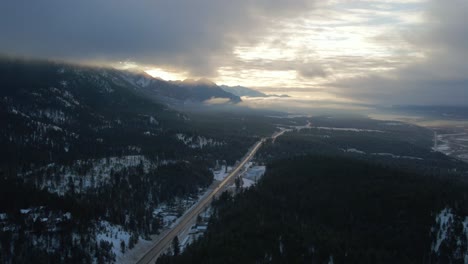 Espléndida-Toma-Aérea-De-La-Autopista-95-Del-Cariboo-Que-Corre-Hacia-El-Horizonte-Rodeada-Por-Una-Gran-Cordillera-Cubierta-De-Nubes-Durante-La-Cálida-Luz-Del-Amanecer:-Vista-Panorámica-Del-Paisaje-Invernal