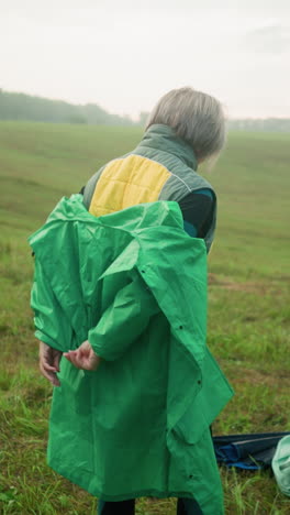 back view of woman wearing yellow and green jacket as she removes green raincoat to pack it on grassy field, umbrella and bag are on the ground, trees in the distance under cloudy sky