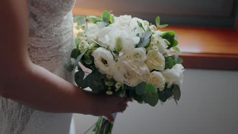 Novia-Sosteniendo-Un-Exuberante-Ramo-De-Flores-Blancas-Junto-A-Una-Ventana,-Mostrando-La-Belleza-De-La-Boda