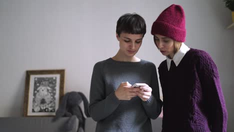 Two-stylish-woman-using-mobile-phone-in-home-interior.-Female-friends-smartphone