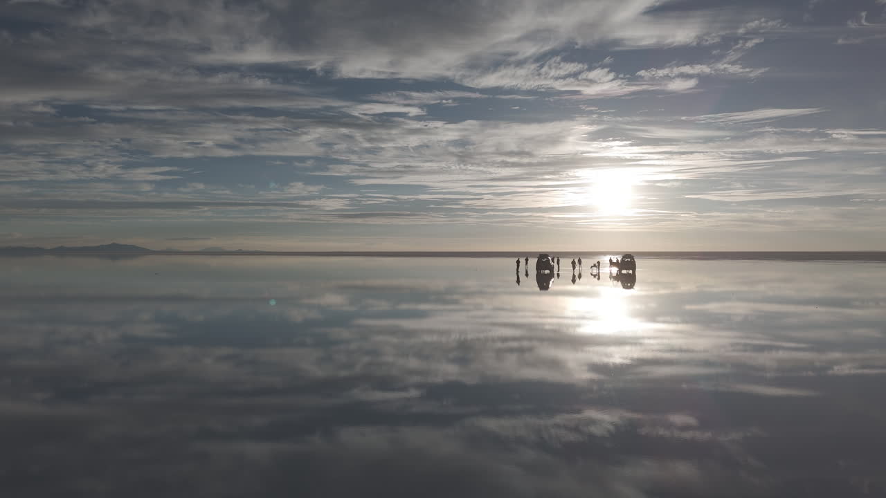 Drone shot flying over the salt flats in Uyuni Bolivia during sunset with reflection in the water and a truck and tourists watching the sun LOG