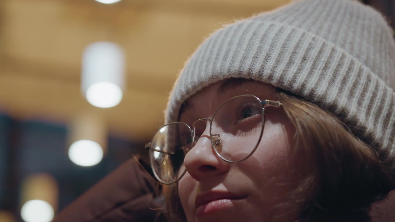 Witte vrouw draagt gebreide muts, ronde bril en zachte woltextuur, close-ups van zijprofiel met warme bokeh-lichten, rustige bedachtzame stemming, late avond student-dichter verzonken in gedachten in stadscafé.