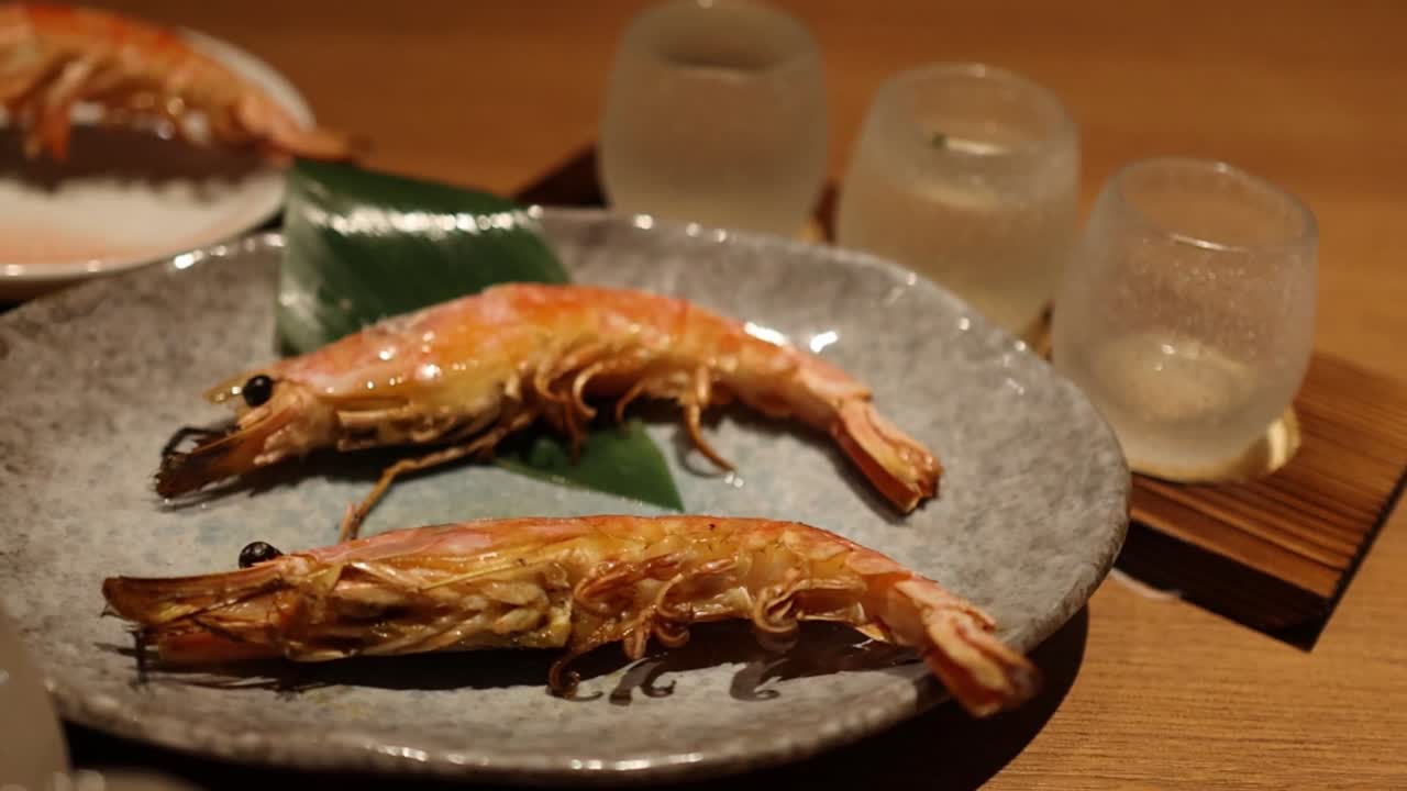 Close-up of chopsticks picking grilled shrimp from a stone plate with leaf garnish and glass cups.