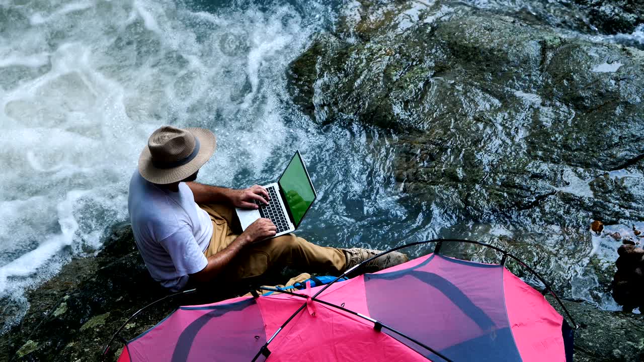 joven usando una computadora portátil de pantalla verde en una cascada y acampando. concepto de viaje y trabajo independiente