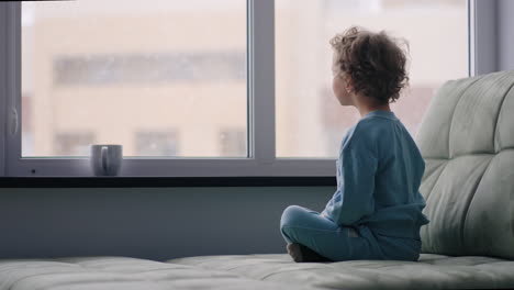 cute curly toddler is looking out window at snowy weather in winter day sitting at home
