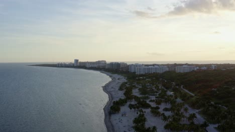 Left-trucking-aerial-drone-shot-of-the-beautiful-tropical-beach-coastline-surrounded-by-palm-trees-and-resorts-on-Crandon-Park-in-Key-Biscayne-outside-of-Miami,-Florida-on-a-warm-sunny-summer-evening