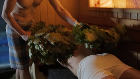 man lying on wooden bench in spa with masseur doing massage with oak broom. client in finn sauna indoors with professional.