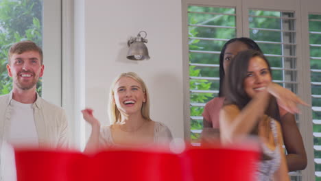 multi-cultural group of friends playing game at home together throwing ball into paper cup