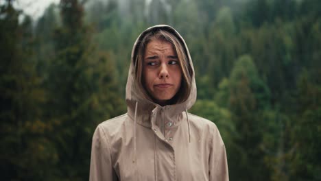 A-sad-blonde-girl-stands-against-the-background-of-forest-rain-in-the-mountains,-she-is-sad-and-looks-at-the-camera