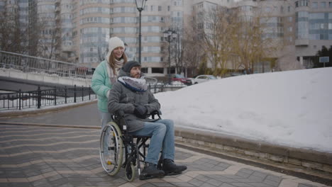 happy caucasian woman taking her disabled friend in wheelchair for a walk in the city and talking together