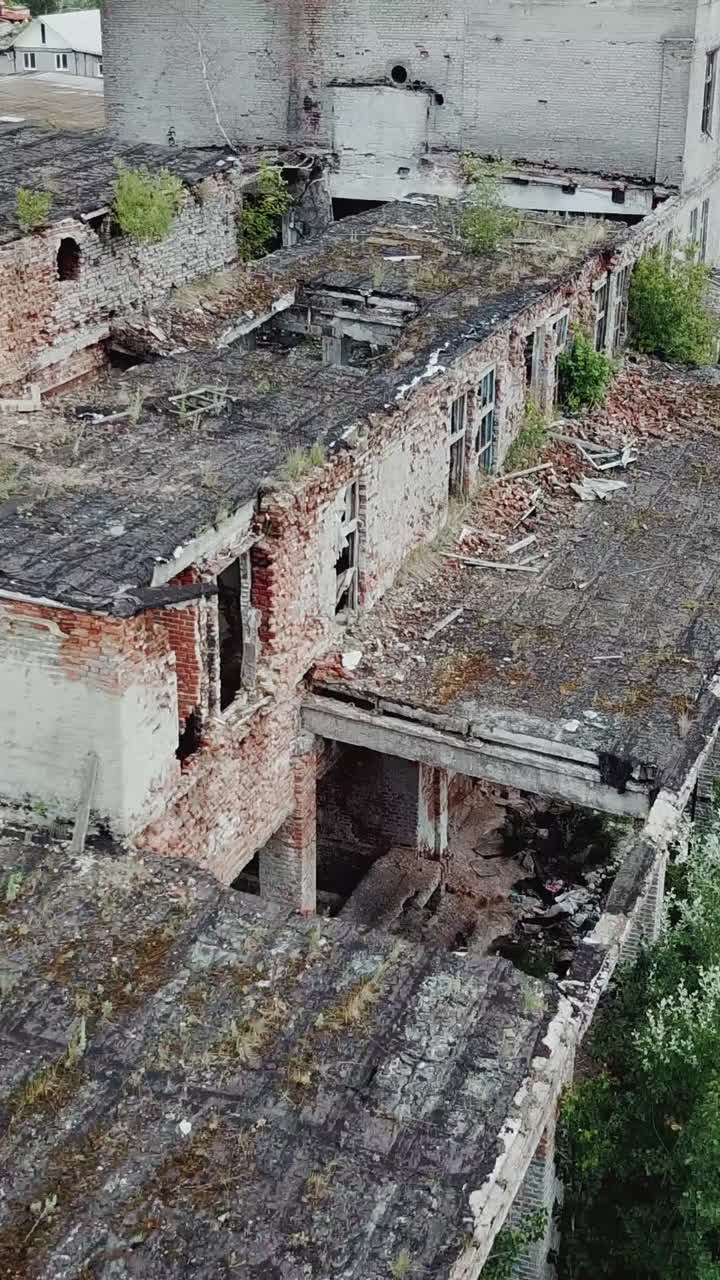 Aerial view. Ruined old building, tilt down to the dry grass and bushes. Abandoned factory halls with broken glass on windows. Apocalyptic scene with destroyed warehouse. Vertical video