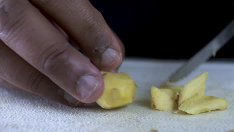 hand of ethnic minority adult male using serrated knife to slice pieces of ginger on cutting board