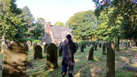 following male wearing hat and casual clothing walking alone in sunlit cemetery towards old chapel