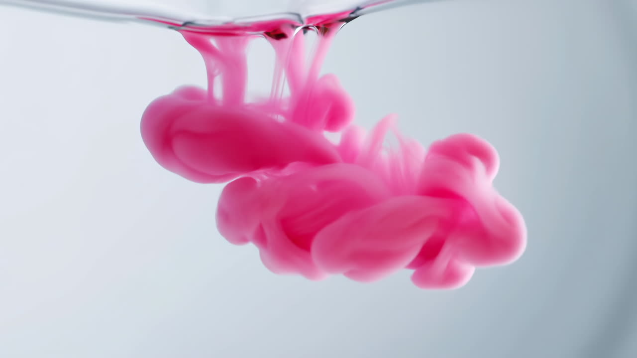 Lyserødt blæk falder ned i vand, et filmisk makrostudie i slowmotion af en hvirvlende fane, med blød studiebelysning.
