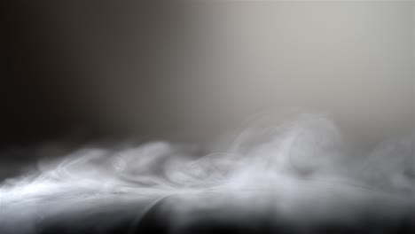 Wispy-strands-of-white-fog-smoke-twirl-move-in-slow-motion-across-black-leather,-grey-background