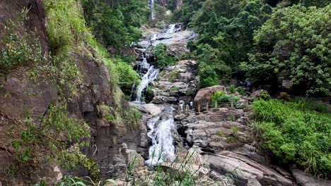 beautiful ravana waterfall cascading over rocks surrounded by lush greenery in ella, sri lanka. a tranquil scene with visitors exploring the natural beauty of the area.