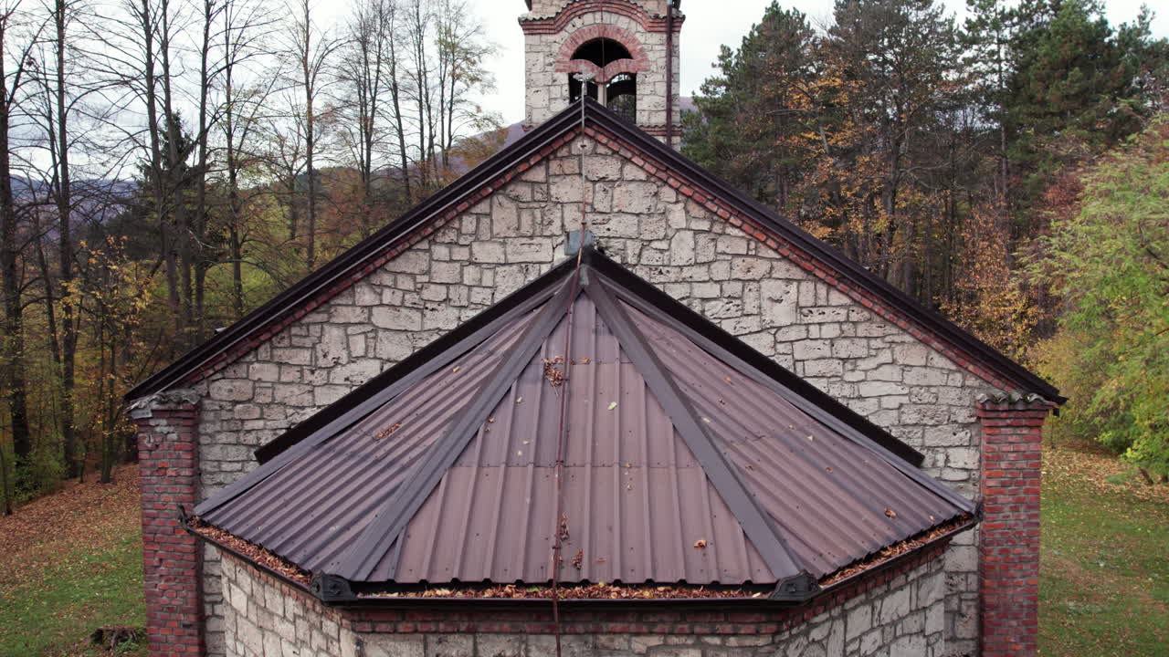 Drone view of the Orthodox church bell tower rising above the roof and autumn woodland in Turbe. Stone and brick details stand out against colorful foliage and mountain scenery
