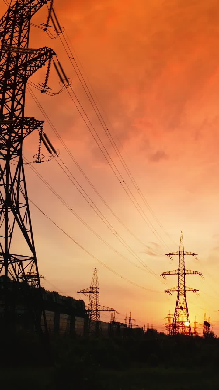 High voltage cable and pylons at sunset. Silhouettes of electric towers against dramatic sky with red setting sun. Vertical video