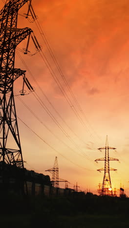 high voltage cable and pylons at sunset. silhouettes of electric towers against dramatic sky with red setting sun. vertical video