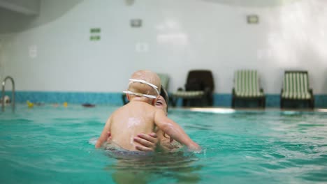 young mother lifting her little boy in protective glasses from the water while teaching him how to swim in the swimming pool