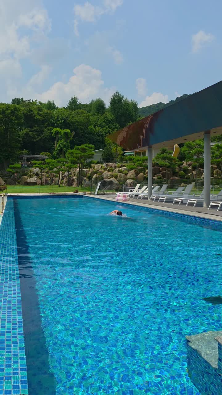 A man swims the crawl stroke in a crystal-clear outdoor pool, captured in a vertical shot with vibrant blue water and lush green landscape in the background