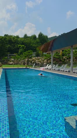 a man swims the crawl stroke in a crystal-clear outdoor pool, captured in a vertical shot with vibrant blue water and lush green landscape in the background