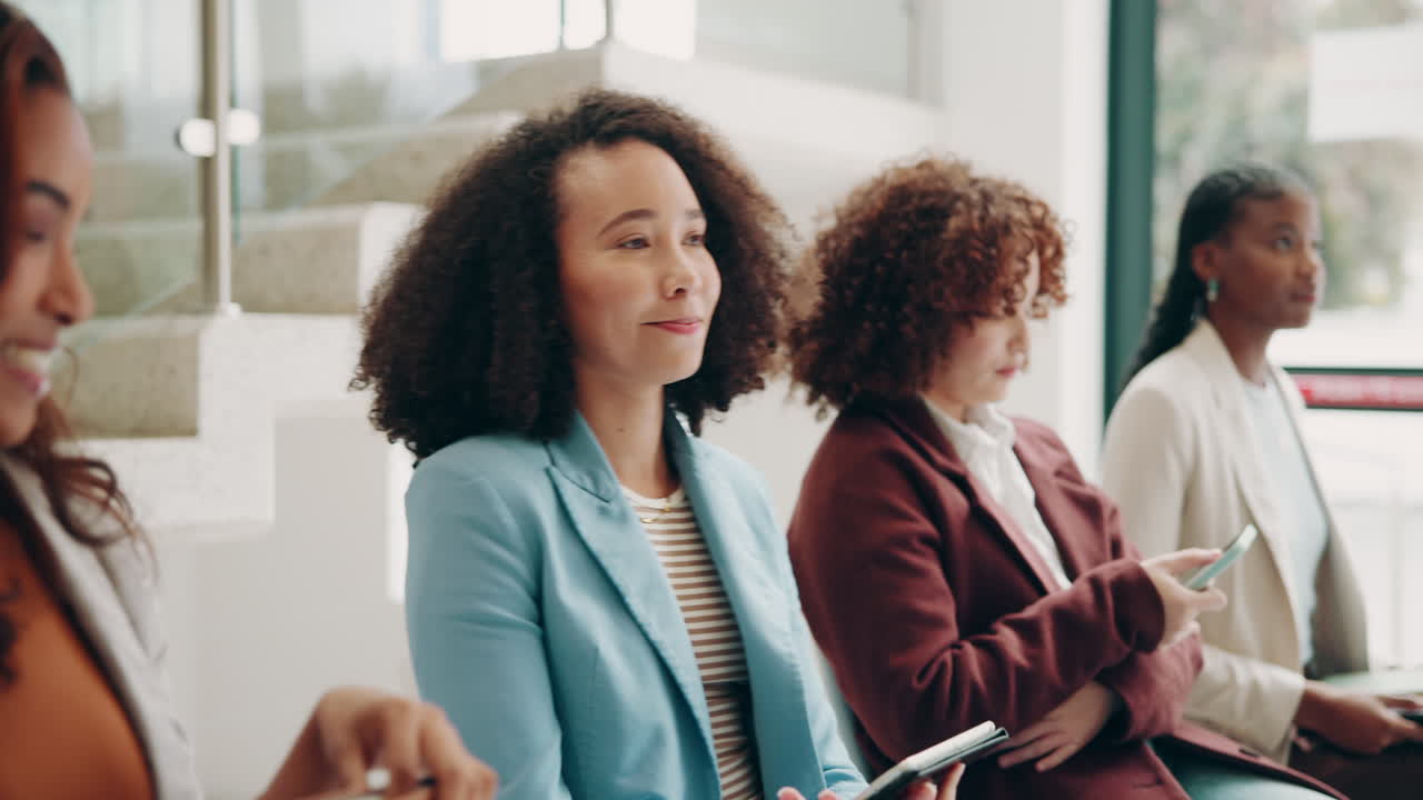 A group of diverse businesswomen sit in a meeting room, listening to a presentation and smiling.