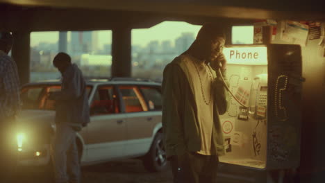 black man talking on street payphone during evening meeting with friends