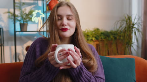 pretty smiling young woman drinking a cup of coffee or herbal tea sitting at home couch break time