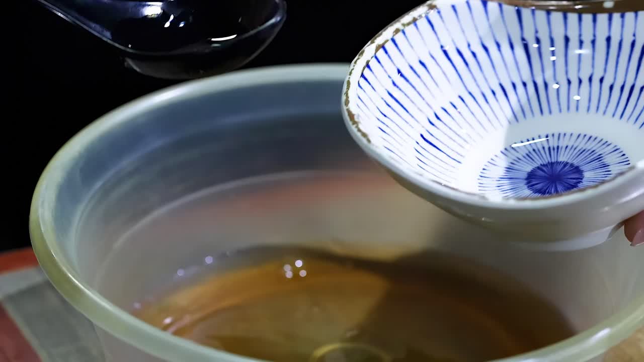A person uses a spoon to pour dark sauce from a small bowl into a larger bowl.