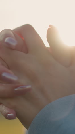 primer plano de una mujer con uñas pulidas realizando una torsión de la muñeca en un suéter azul, la luz del sol creando un brillo cálido alrededor de las manos, destacando el movimiento y la elegancia en un entorno al aire libre sereno