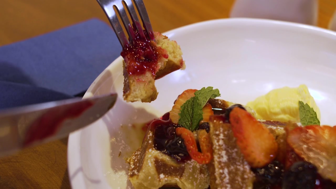 Persona che taglia un waffle belga dorato con forchetta e coltello, in un ambiente moderno da brunch in caffetteria.