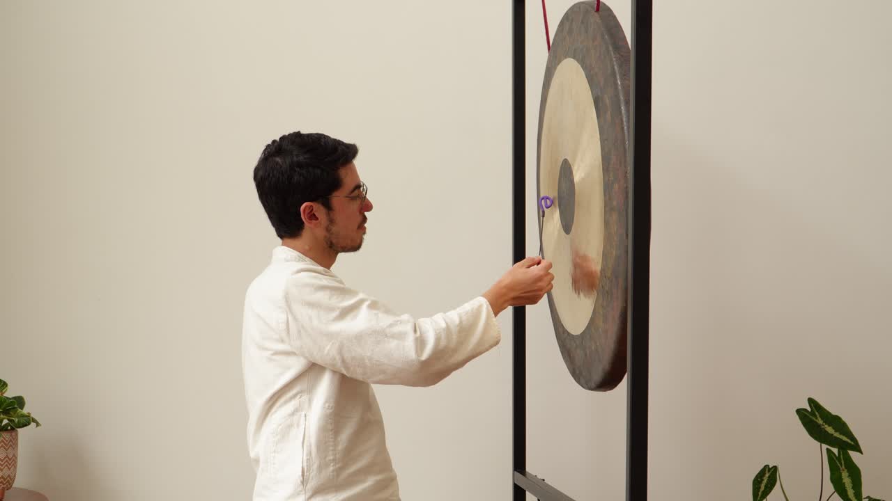 Side view of a man rubbing a large symphonic gong with silicone friction mallets to produce sustained singing tones in a calm minimalist indoor studio—ideal for meditation, sound healing and wellness