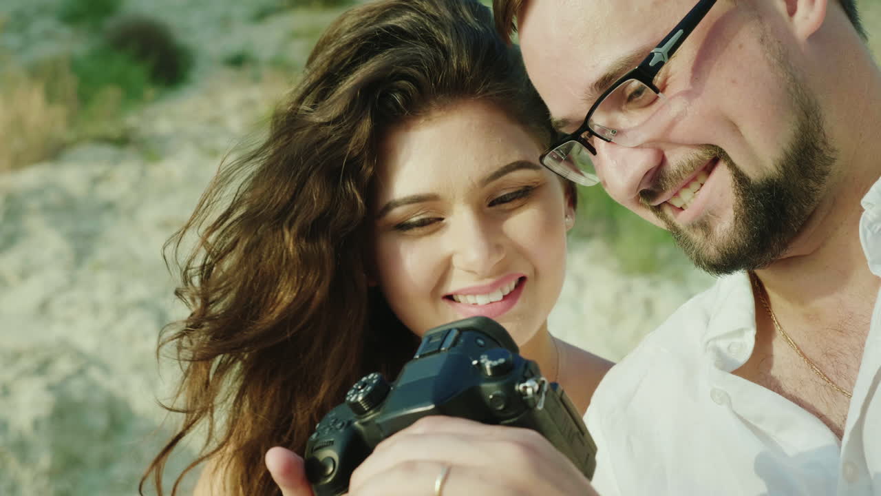 Um jovem casal olha as fotos em uma câmera digital, sentados perto da praia. Grandes lembranças.