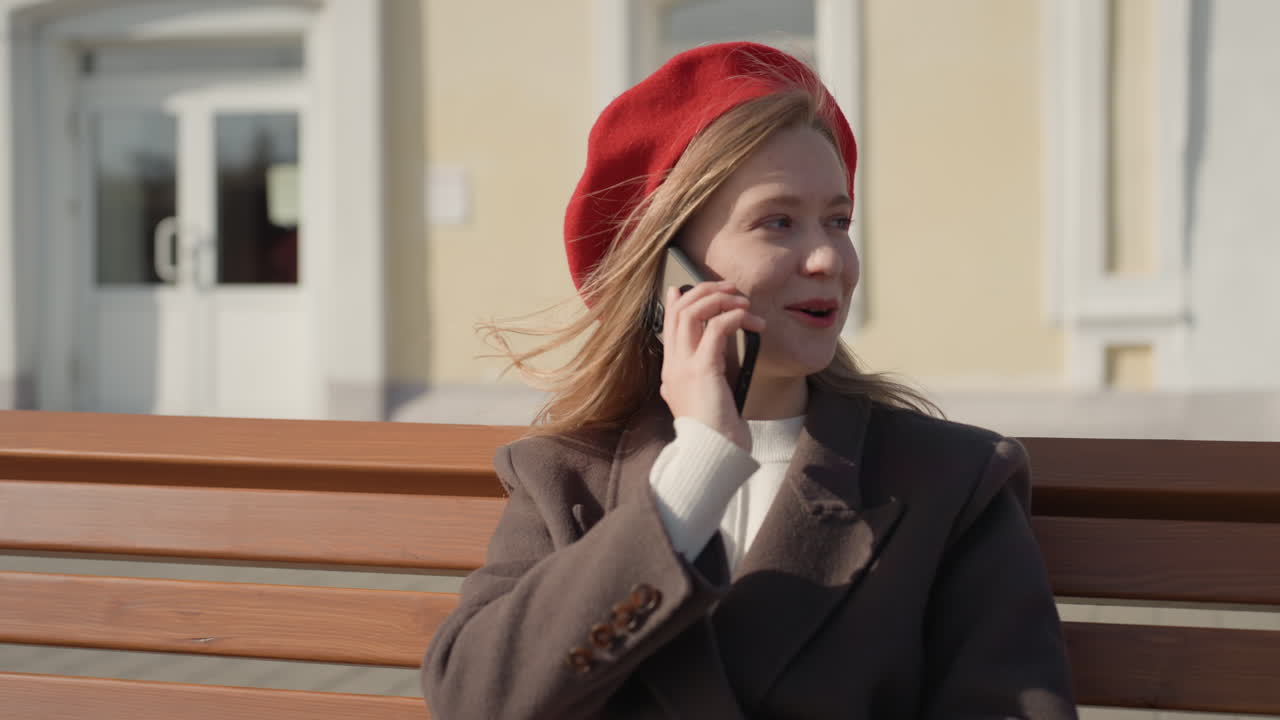 Donna caucasica su panchina che parla, atmosfera da studentessa e scrittrice freelance che controlla l'agenda mentre si gode un parco urbano soleggiato. Cappotto caldo, berretto rosso, postura rilassata, sorriso spontaneo e leggera brezza. Vetrina sullo sfondo.