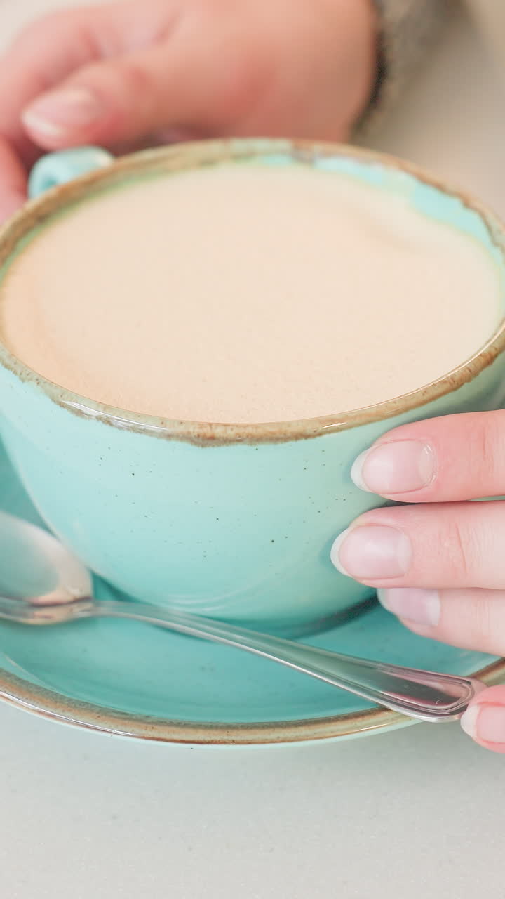 manos caucásicas levantando una taza turquesa de su plato, cliente terminando un latte en una cafetería, vista desde arriba que revela un plato y una cuchara turquesas vacíos, restos de espuma suave en el borde, tonos interiores pastel, ordenado
