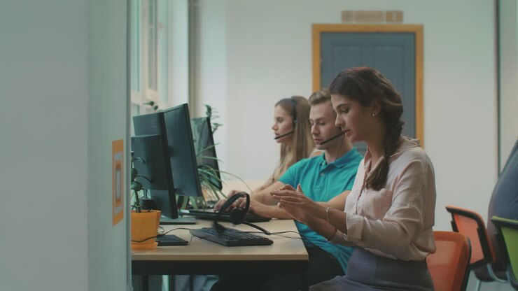 Mladá žena hovoří s klienty v coworkingovém prostoru. Unavený pracovník odhazuje sluchátka.