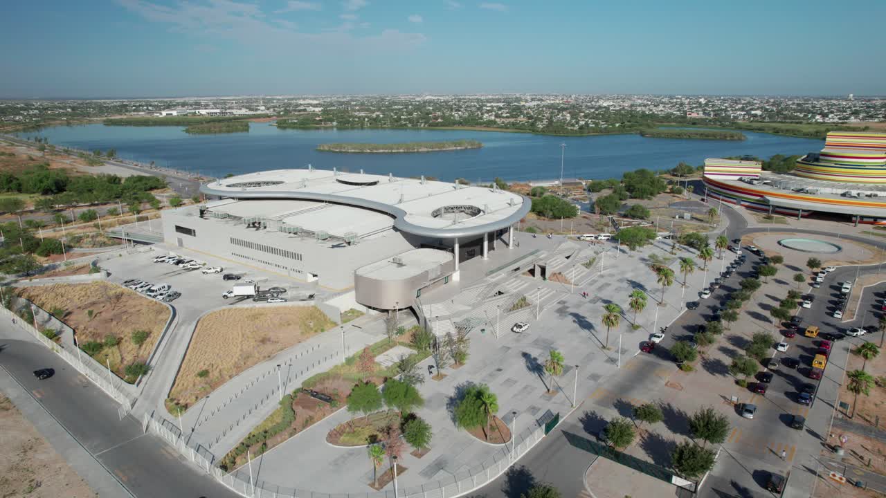 Aerial view of a modern architectural building complex situated by a lake with a distant cityscape.