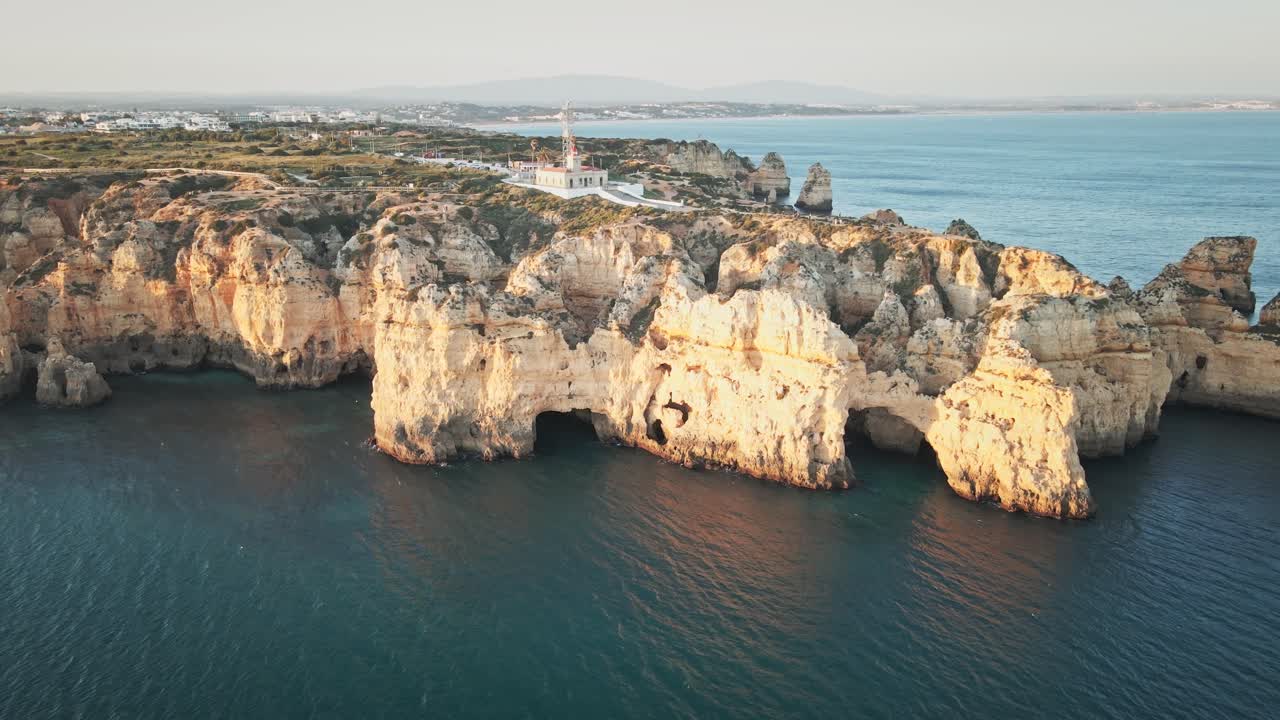 Camera moves forward, sweeping across sunlit limestone cliffs and arches, and gliding toward Ponta da Piedade lighthouse, with the coastline and town on the horizon