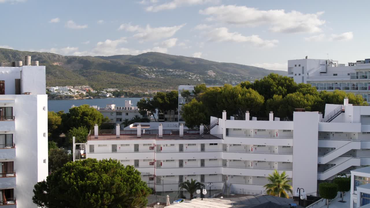 ручной снимок отелей магалуфа, пышных деревьев и прибрежных холмов с спокойным видом на залив под частично облачным небом.