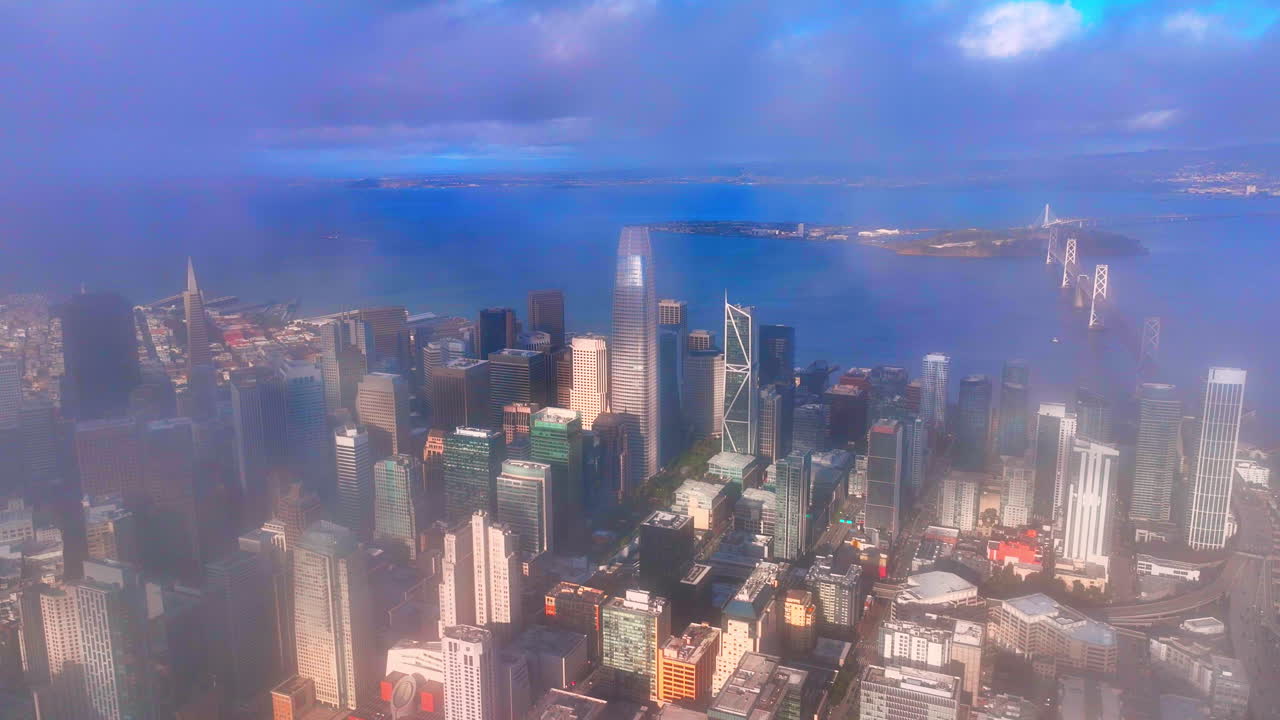 High-angle aerial view of the downtown San Francisco financial district, featuring high-rise buildings and the Bay Bridge partially obscured by morning fog