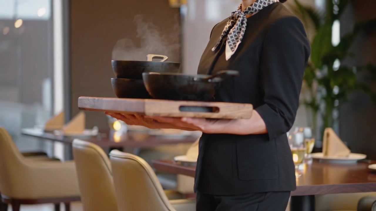Server entering in black uniform carrying tray with steaming iron pans in dining room, serving