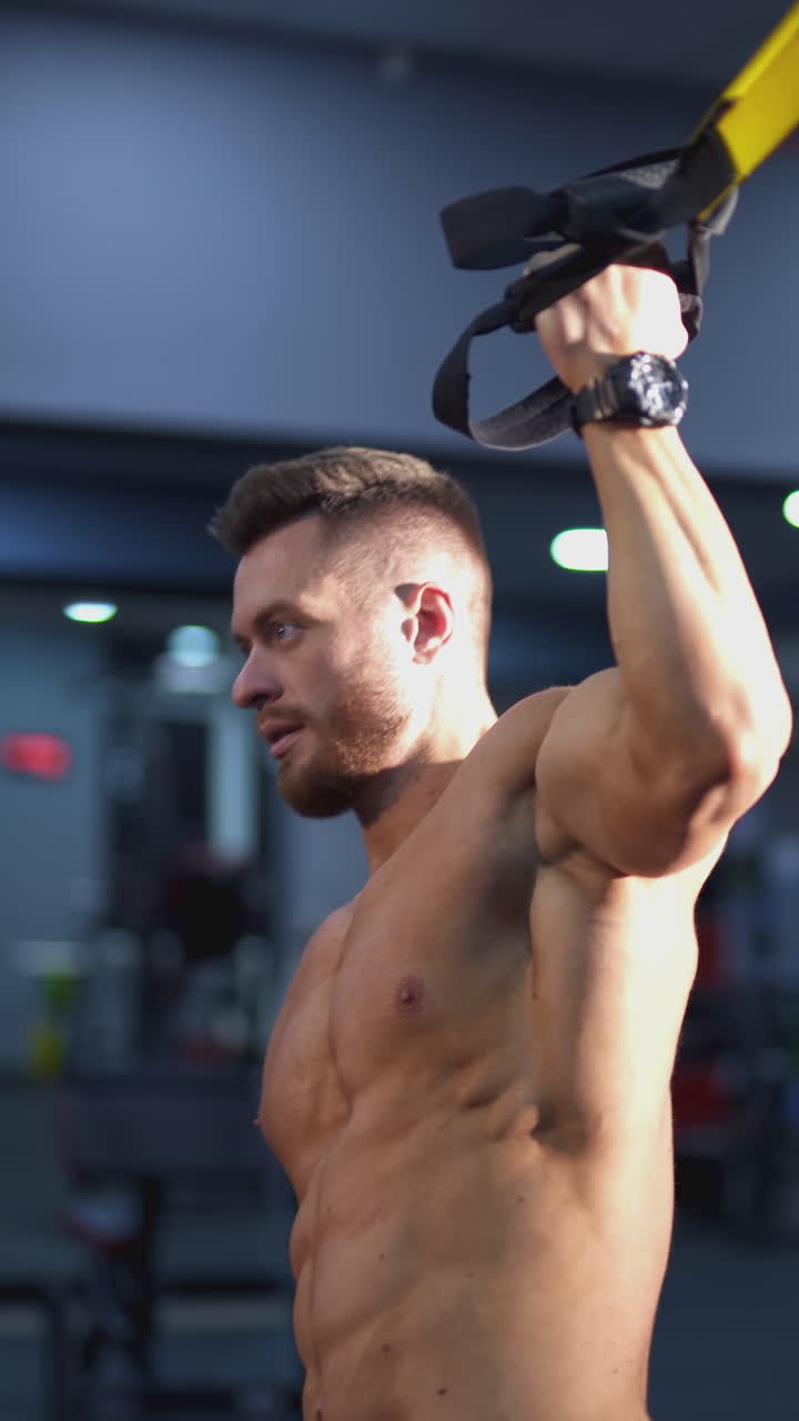 Concentrated sportsman tilts his body to the side. Muscular man trains near the simulator in fitness center. Strong athlete doing exercise indoors. Side view. Vertical video