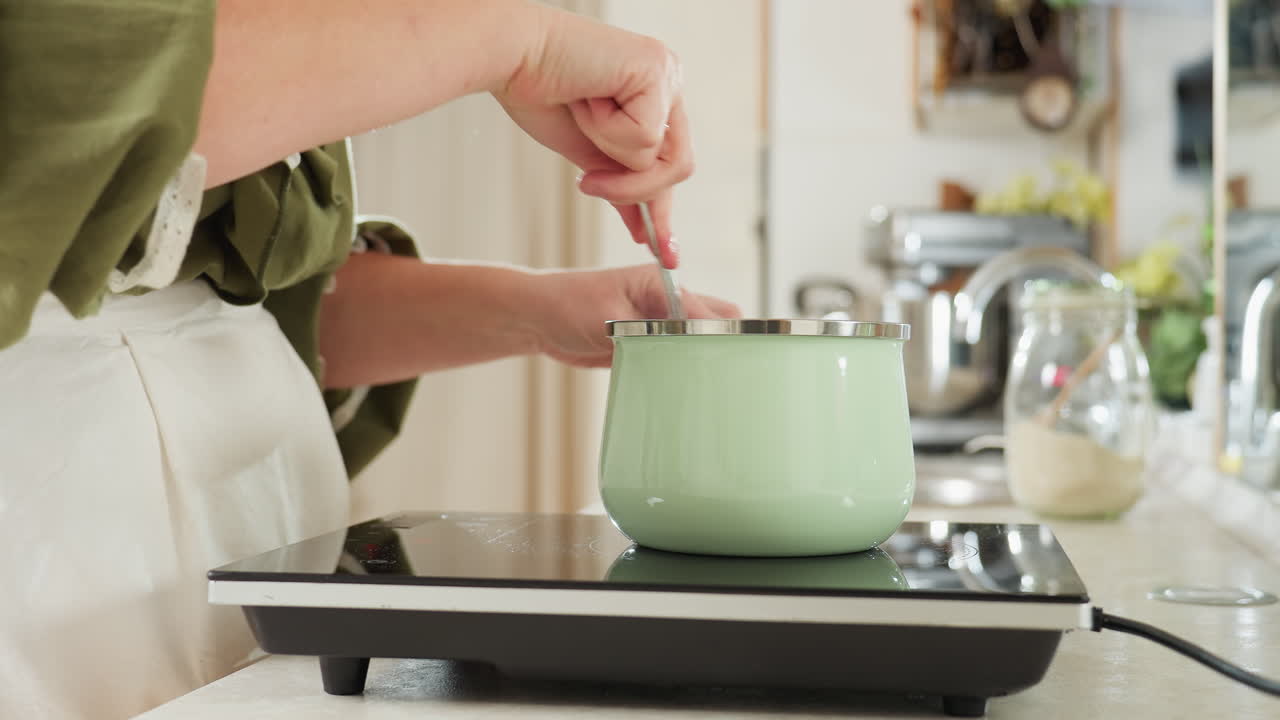 Woman in green blouse and white apron stirs food in pastel green pot on electric stove in bright kitchen, preparing meal with care while surrounded by modern appliances