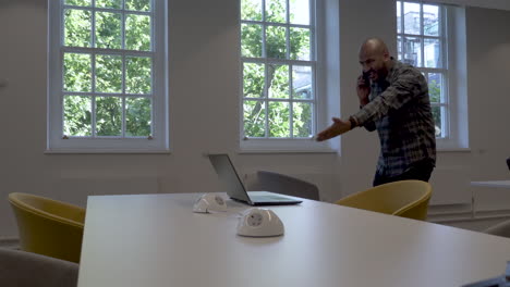 an asian indian man pacing around an office while talking on his mobile phone, the man expressing his anger and frustration while gesturing and pointing to the screen of his laptop computer