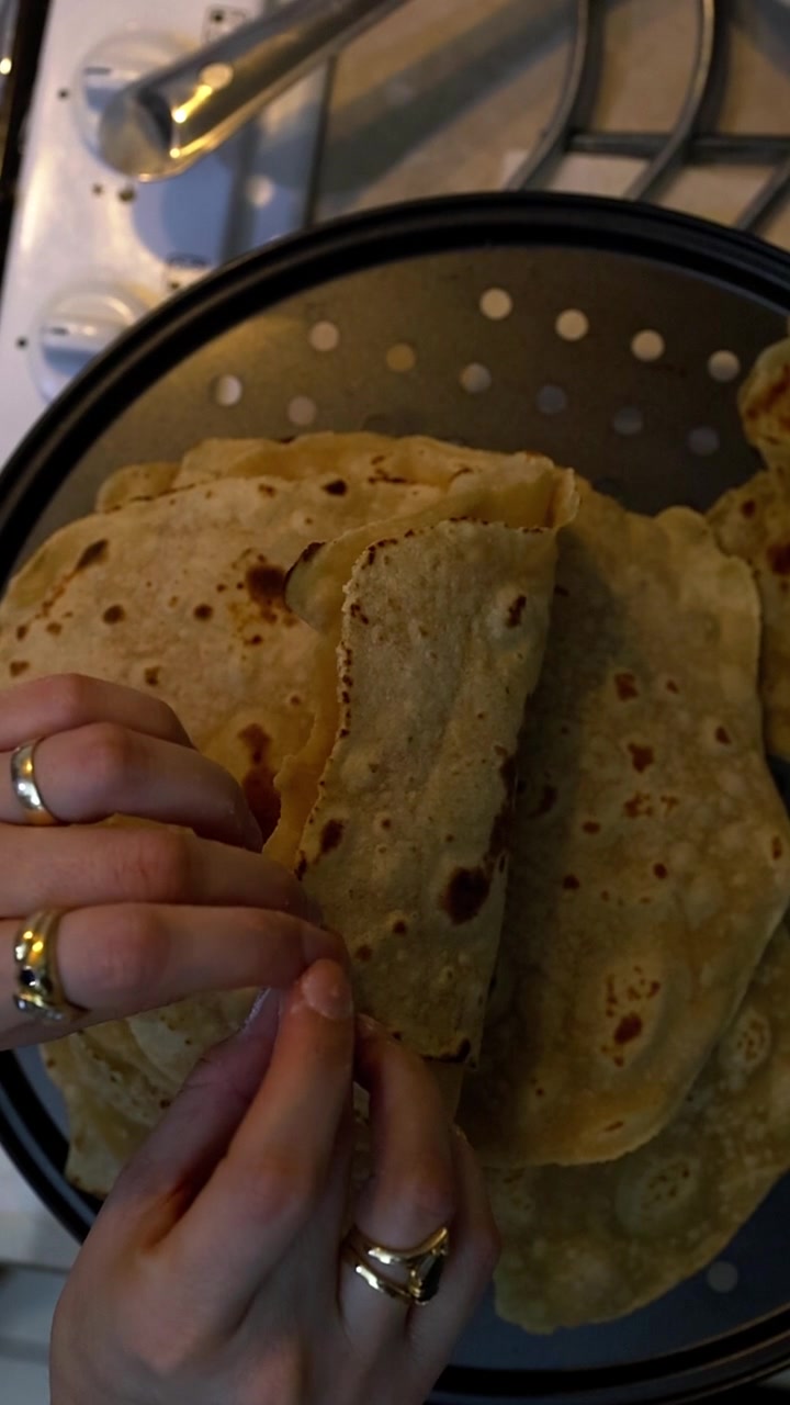 Close-up tangan mengangkat roti pipih chapati hangat berwarna keemasan dari panci kukus berlubang hitam di atas kompor gas. Roti tanpa ragi berwarna cokelat keemasan dengan bintik-bintik gosong yang terlihat, masakan tradisional India