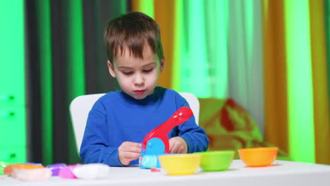 focused baby boy in blue sweater sits at desk busy with plasticine. lovely child is busy with his hobby at home.