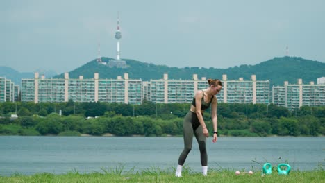 젊은 스포츠 여성이 강변의 한강 공원에서 몸의 근육을 따뜻하게 하고 긴장시 ⁇ 니다. 배경에는 서울 남산 타워 전망이 있습니다.