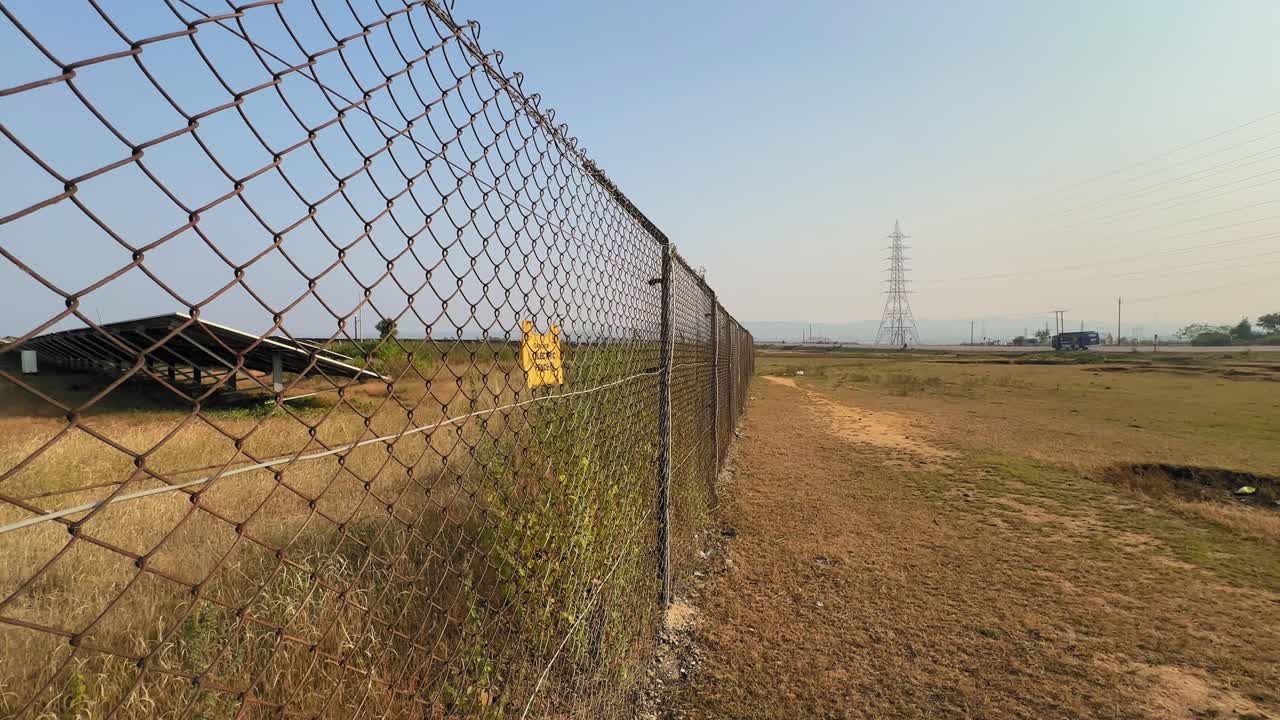 Tracking shot along an electric fence beside a solar farm, showing metal mesh, warning sign, dry grassland, and distant power towers under clear sky